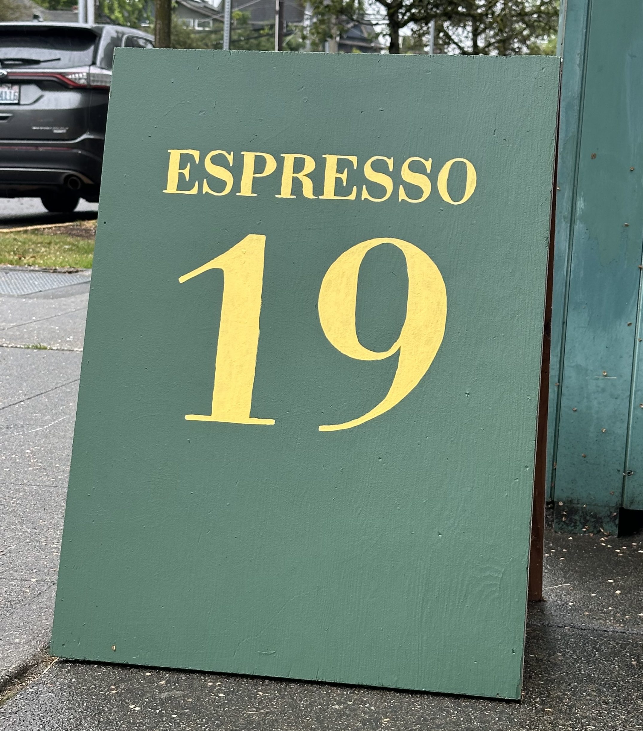 A painted pavement sign, also called sandwich board, all dark green with espresso 19 in yellow lettering.